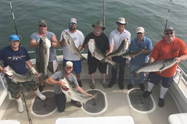 View from atop the boat looking down at the deck at 8 men each smiling for the camera and holding a striped bass.