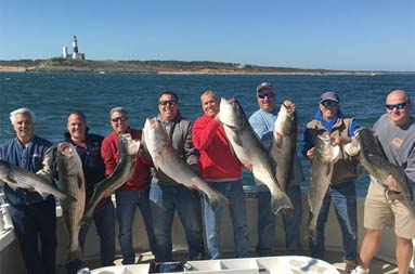 With a clear blue sky and the lighthouse in the background, 8 men smile and proudly hold up their striped bass.