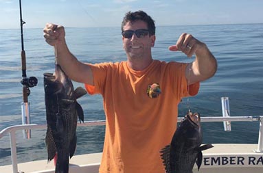 A man wearing an orange t-shirt and sunglasses holds up his line with two 2 sea bass hooked on it.