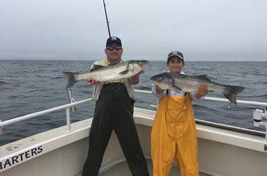 A father, wearing black waders, stands next to his teenage son wearing yellow waders, each holding up a striped bass.