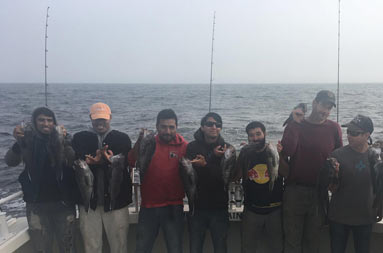 On a gray overcast day, nine men smile as they each hold up the 2 sea bass they caught.