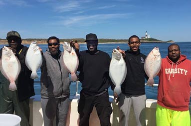With a sunny blue sky, calm waters and Montauk lighthouse in the background, a group of 5 men each smile and hold up 2 fluke.