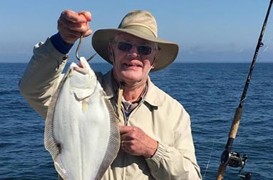 A 80 yr old man wearing a hat smiles as he holds up his fluke.