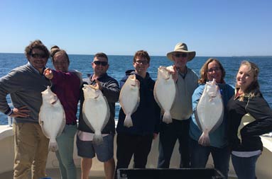 A family of 7, including the guest of honor, Wilson whose birthday it was, smile on the boat together and hold up some of the fluke they caught.