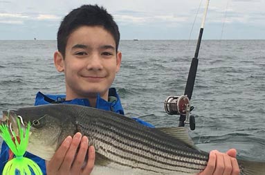 An adolescent boy wearing a blue jacket smiles as he holds up his striped bass with 2 hands.