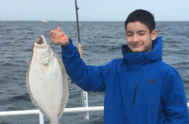 An adolescent boy wearing a blue jacket smiles as he holds up a fluke still on the line.