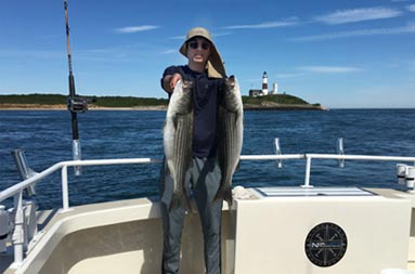 With a  bright blue sky in the background and the lighthouse behind him, a young adult holds up 2 striped bass.