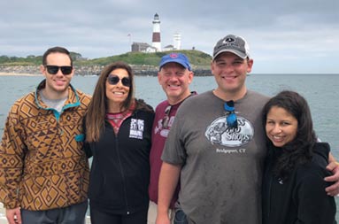 A mother and father, along with 2 sons and and a daughter, smile for the camera, with the lighthouse behind them in an overcast sky.