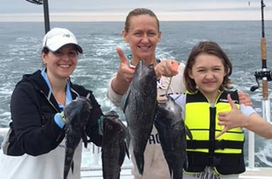 Two adult women and an adolescent girl each hold up sea bass as they smile for the camera.