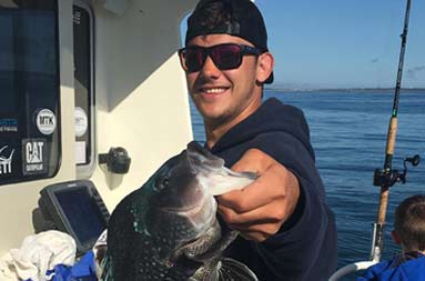 On a sunny day, a guy smiles and holds out the sea bass he caught.
