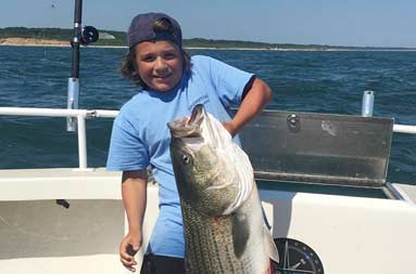 An adolescent boy holds up a large striped bass with one hand and smiles for the camera.