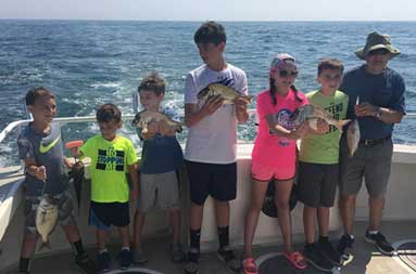 With the sunny sky and glistening ocean behind them, seven kids ranging in age, and an adult, each hold up the porgy they caught.
