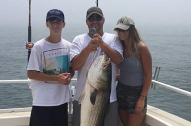 A man smiles and holds up a large striped bass with two teenagers, boy and girl, on either side of him.