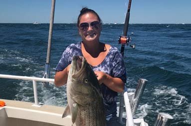On a bright sunny day, a woman smiles and holds up a large striped bass with two hands.