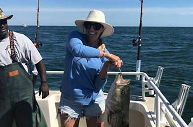 With First Mate Gil looking on, a woman uses two hands to hold up her large striped bass for the camera.
