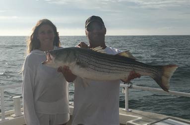 With the sun casting a shadow on them from behind, a man and woman stand next to one another holding up a striped bass.