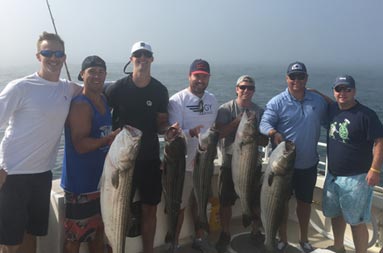 On a very foggy morning, seven men pose for the camera as some hold up the striped bass they caught.