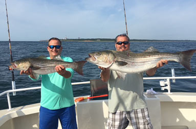 Two men wearing sunglasses smile and each hold out their striped bass for the camera.