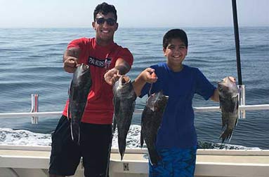 A young man and adolescent male smile and hold up the sea bass they caught.