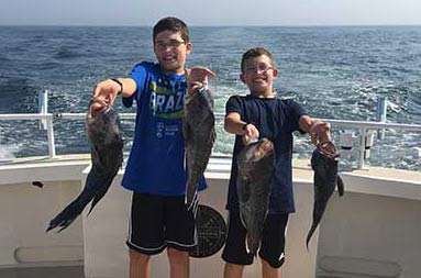 Two adolescent boys smile big and hold up two sea bass each.