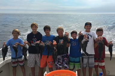 Seven adolescent boys stand together as each hold up 2 sea bass for the camera and smile.