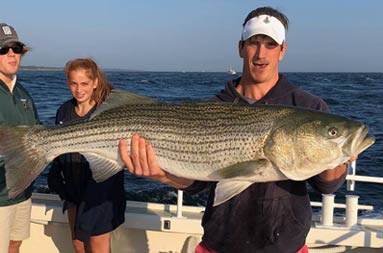 A man holds up a striped bass with  2 people standing in the background.