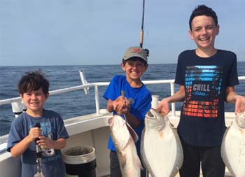 Three boys smile big as they hold up the fish they caught.The youngest holds up porgy (scup), the middle holds up striped bass, and the eldest holds up 2 fluke.