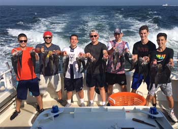 Seven guys each hold up 2 sea bass and smile for the camera on a sunny day on the water.