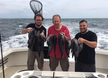 With an overcast, but slightly sunny sky, behind them, three men each hold up 2 sea bass for the camera.