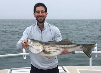 With a grayish/blue sky in the background, a man proudly holds up the striped bass he caught and smiles.