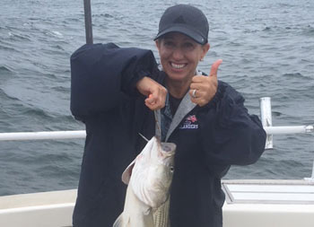 A woman wearing a blue jacket and baseball cap, gives a thumbs up to the camera as she holds up her striped bass with the other hand.