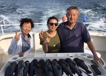 With the wake of the boat behind them, two women and a man smile for the camera and give the peace sign, while they stand behind the field table with a spread of sea bass.
