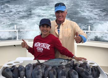 With the wake of the boat trailing behind, a man wearing a yellow shirt and blue baseball cap, and a woman wearing a red Montauk sweatshirt, give thumbs up in front of a pile of sea bass laid out on the filet table.