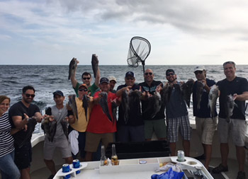 A group of one woman and 11 men smile for the camera as some hold up the sea bass they caught.