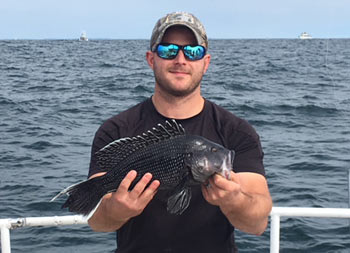 A man wearing a baseball cap and sunglasses holds out the sea bass he caught for the camera.