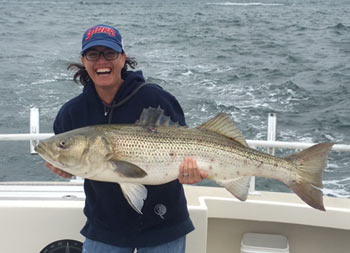 A woman gives a big smile as she holds up a striped bass with two hands.