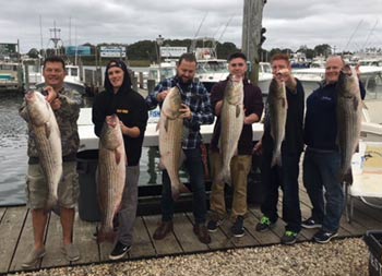 A group of six men stand in a group on the dock, and each holds up the striped bass they caught.