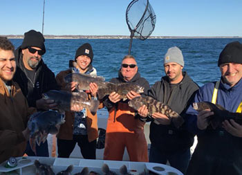 A group of 6 men wearing sweatshirts and hats, stand on the boat on a crisp fall day with blue sky behind them, holding up the fish they caught.