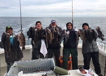 One woman and four men wearing winter coats and hats each smile and hold up the fish they caught.