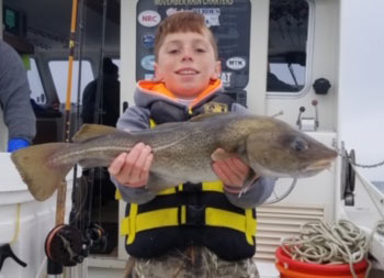 Photo of an adolescent red-haired boy wearing a life jacket and standing on the boat proudly holding up the cod he caught.