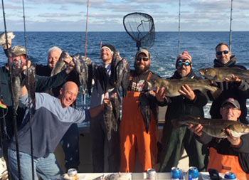 On a cold fall day on the water, a group of 8 men smile for the camera and hold up the fish they caught.
