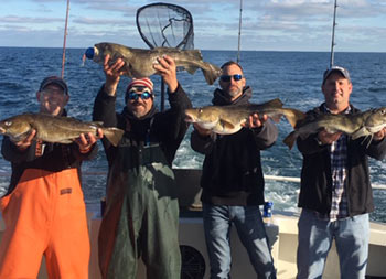 Four men wearing sweatshirts and hats in the cold sunny day, smile for the camera and hold up the fish they caught.