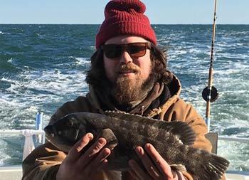On a cold day, a man wearing a hat and coat, holds up a blackfish (tautog).