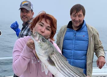 A woman with short red hair kisses the striped bass she caught as a man behind her smiles and looks on.