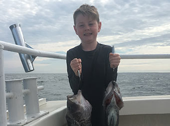 A young boy smiles and uses his strength to hold up a sea bass in each hand.