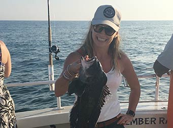 With sunset drawing near, a woman wearing shorts and sunglasses, holds up sea bass and smiles.