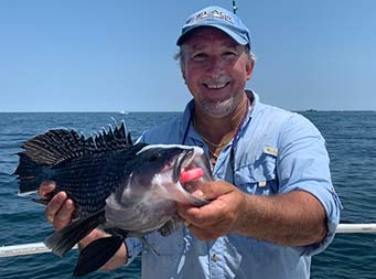 With the blue sunny sky and ocean behind him, a man smiles and holds up the sea bass he caught.