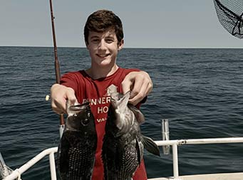 A young man wearing a dark red tshirt holds out the 2 sea bass he caught for the camera .