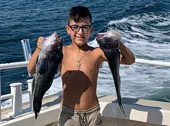 A young boy wearing glasses smiles and proudly holds up a sea bass in each hand.