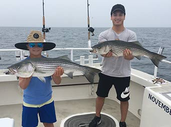 On an overcast day, a young boy and man both hold up a striped bass and smile for the camera.